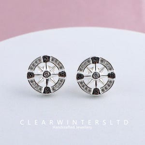 Puede incluir: Un par de pendientes de botón plateados. Cada pendiente presenta un diseño de brújula con los puntos cardinales (N, O, E, S). La brújula está rodeada de pequeñas piedras transparentes. El texto "CLEARWINTERSLTD Handcrafted Jewellery" está en la parte inferior.
