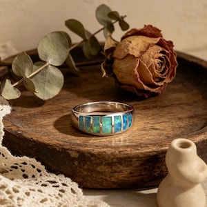 Peut inclure: Une bague en argent ornée d'une bande de pierres rectangulaires bleu et vert iridescentes. La bague est posée sur un plateau en bois avec une rose séchée et des feuilles d'eucalyptus. Le design de la bague est une version moderne d'un style classique.