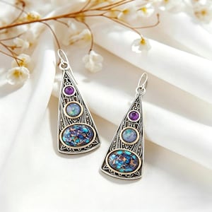 Può includere: Un paio di orecchini triangolari color argento con dettagli intricati. Ogni orecchino presenta tre pietre incastonate: una viola, una azzurra e una grande pietra ovale con motivi blu e turchese, incastonata in una montatura dorata.