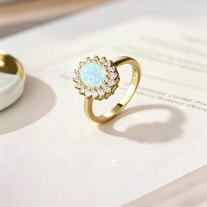 May include: A gold ring with an oval opal gemstone at its centre, encircled by a halo of smaller, clear stones. The ring's band is plain gold. The opal has a pale blue colour, and the ring is set against a white background.