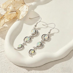 May include: A pair of silver earrings with a drop design. Each earring features three oval-shaped opals set in a swirling silver frame. The opals have a milky white and iridescent appearance.