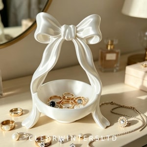 May include: A white ceramic jewellery dish featuring a sculpted bow design at the top. The bowl base holds several gold-toned rings and small jewellery pieces. Additional rings and a delicate chain necklace are arranged around the base of the stand.