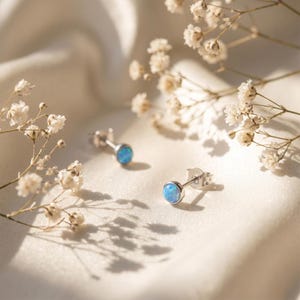 May include: Two silver stud earrings, each featuring a round, pale blue opal. The earrings are set against a backdrop of white, flowing fabric and small white flowers. A simple and elegant jewellery design.