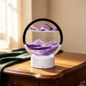 May include: A circular sand art display featuring layers of purple and white sand forming mountain-like shapes inside a clear glass frame. The frame is mounted on a solid white cylindrical base with a small power button and indicator lights.