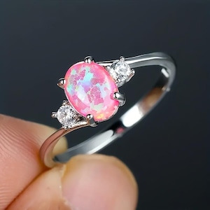 May include: A silver ring with a large, oval pink opal gemstone, displaying iridescent colours. Two smaller, clear gemstones are set either side of the opal. The ring has a slender, polished band.