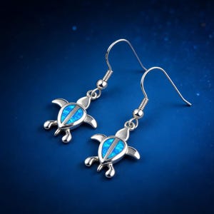Könnte beinhalten: Ein Paar silberfarbene Ohrringe mit einem Schildkrötendesign. Jeder Ohrring hat eine blaue, opalartige Mitte und detaillierte Flossen und Kopf. Die Ohrringe hängen an einem einfachen Haken.