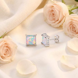 May include: A pair of square opal stud earrings with a silver setting. The opals display a mix of colors, including pink, blue, and yellow. The earrings are set against a soft, white background with pink roses and petals.