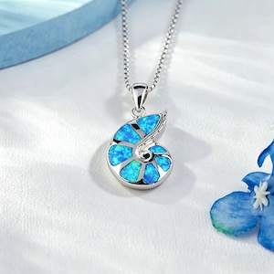 Puede incluir: Un collar plateado con un colgante en forma de concha de nautilus. La concha está rellena de ópalo azul brillante, creando un contraste con la plata. Este delicado collar es una joya.