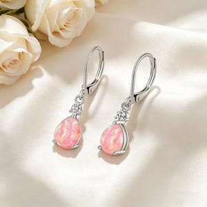 May include: A pair of silver drop earrings with a pear-shaped, pink opal stone. Each earring features a small, round, clear gemstone above the opal. The earrings are displayed on a white, satin-like surface.