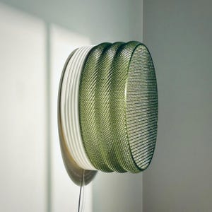 Puede incluir: Una moderna lámpara de pared con un diseño en capas. Presenta anillos concéntricos en blanco y verde, con una textura de malla en los anillos exteriores. La lámpara está montada en una pared y tiene un cable de alimentación.