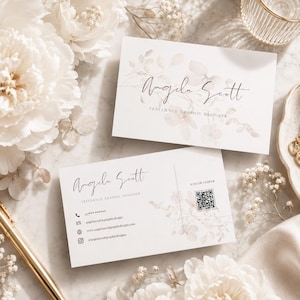 Op de afbeelding: Twee witte visitekaartjes met de naam "Angela Scott" en "Freelance Graphic Designer" gedrukt in zwart script. Bloemenontwerpen accentueren de kaarten. Eén kaart bevat een QR-code en contactgegevens. De kaarten zijn omgeven door witte bloemen en een gouden pen.