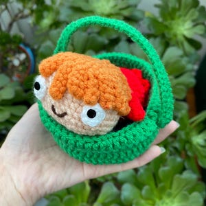Puede incluir: Una cesta de ganchillo verde, con un asa, que contiene un pequeño personaje pelirrojo con una cara sonriente y ojos blancos. El interior de la cesta es rojo. El fondo está lleno de plantas verdes.