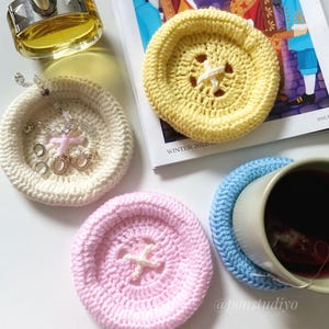 May include: A collection of handmade crochet items, including coasters and small dishes. The coasters are in yellow, pink, and blue, with a star-shaped design in the center. A white dish holds jewelry. A perfume bottle and a magazine are also present.