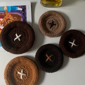 May include: A collection of five handmade crochet coasters in various shades of brown and tan. Each coaster features a button-like design with a contrasting cross stitch in the center. The coasters are arranged on a white surface, with a perfume bottle in the background.
