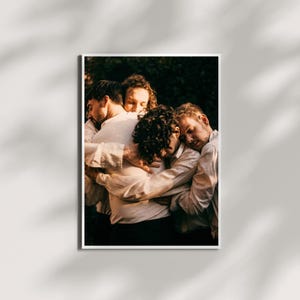 May include: A framed print featuring five people embracing. They are wearing white shirts and dark pants. The image has a warm, natural lighting and a soft focus, creating a sense of intimacy and connection. The background is blurred, suggesting an outdoor setting.