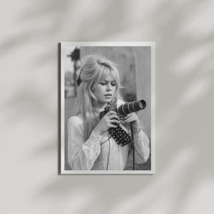 May include: Black and white photograph of a woman holding a vintage camera. The woman has long, wavy hair and is wearing a light-colored blouse with a polka-dot scarf. The photograph is framed in a white border.