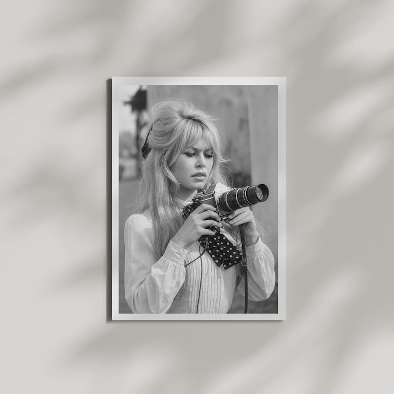 May include: Black and white photograph of a woman holding a vintage camera. The woman has long, wavy hair and is wearing a light-colored blouse with a polka-dot scarf. The photograph is framed in a white border.