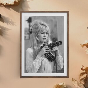 May include: Black and white framed photograph of a woman holding a vintage camera. The woman has blonde hair and is wearing a white blouse with a polka dot scarf. The photograph is in a wooden frame.