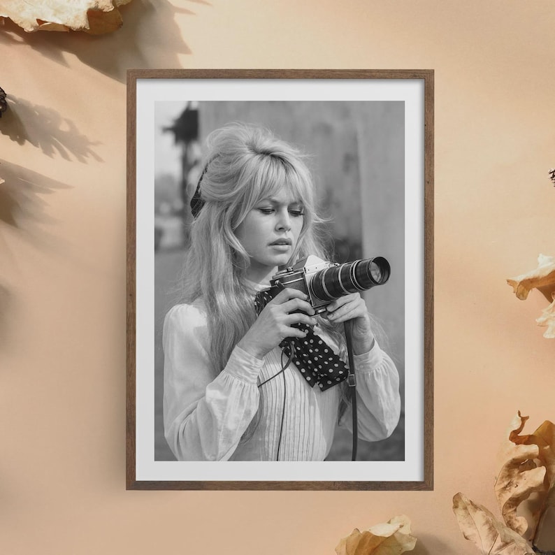 May include: Black and white framed photograph of a woman holding a vintage camera. The woman has blonde hair and is wearing a white blouse with a polka dot scarf. The photograph is in a wooden frame.