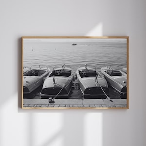May include: Black and white framed photograph of four vintage wooden motorboats docked at a pier. The boats are tied with ropes, and the calm water reflects the sky. A small boat is visible in the distance.