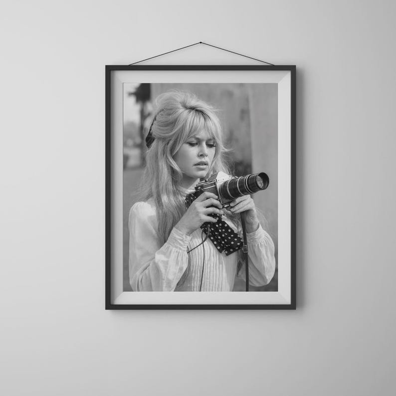 May include: Black and white framed photograph of a woman holding a vintage camera. The woman has long blonde hair and is wearing a light-colored blouse. The camera has a long lens and a polka-dot strap.
