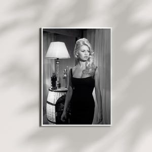 May include: Black and white framed print featuring a woman in a black dress. The image includes a lamp, a decorative table, and bottles. The woman has blonde hair and is looking to the side. The print is displayed on a white wall.