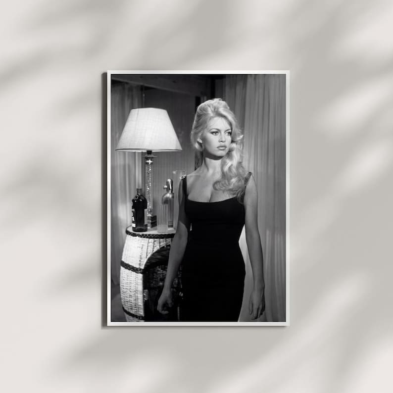 May include: Black and white framed print featuring a woman in a black dress. The image includes a lamp, a decorative table, and bottles. The woman has blonde hair and is looking to the side. The print is displayed on a white wall.