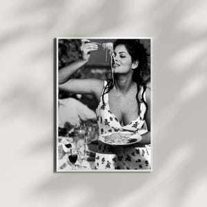 May include: Black and white photograph of a woman enjoying spaghetti. She is wearing a floral print dress and holding a fork with pasta. A plate of pasta and wine glasses are on the table.