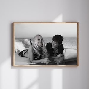 May include: Black and white framed photograph of a couple on a boat. The woman is lying down, smiling, and the man is sitting next to her. The boat is moving on the water, with the ocean and sky in the background.