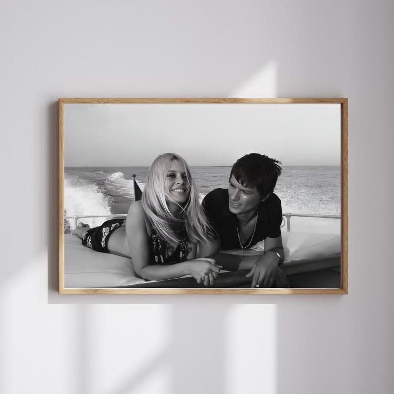 May include: Black and white framed photograph of a couple on a boat. The woman is lying down, smiling, and the man is sitting next to her. The boat is moving on the water, with the ocean and sky in the background.