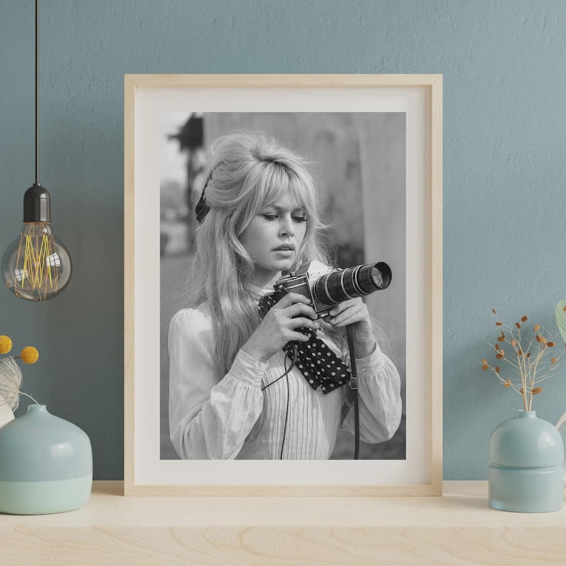 May include: Black and white framed photograph of a woman holding a vintage camera. The woman has long, wavy hair and is wearing a light-colored blouse. The photograph is in a wooden frame and is displayed on a light-colored surface.