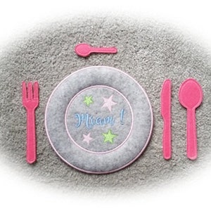 May include: A felt play set for pretend play, featuring a gray plate with a pink rim and the word "Mmm!" embroidered on it, a pink fork, knife, spoon, and a smaller pink spoon.