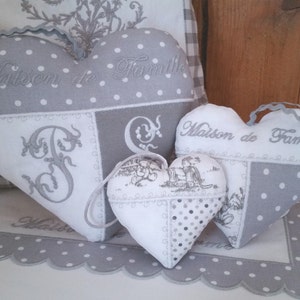 Peut inclure: Trois coussins en forme de cœur gris et blanc avec du texte brodé. Le plus grand coussin porte l'inscription "Maison de Famille" et présente une grande lettre "F" brodée. Le coussin moyen porte l'inscription "Maison de Famille" et le plus petit coussin présente un motif floral avec deux personnages.