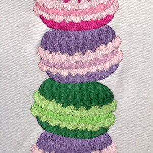 May include: A stack of four colorful macarons with pink, purple, green, and purple frosting. The macarons are arranged vertically, with the pink macaron on top and the purple macaron on the bottom.