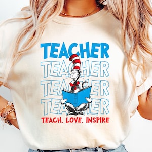 May include: Cream-colored t-shirt featuring the word "TEACHER" stacked four times in blue, with a Cat in the Hat illustration reading a blue book. The text "TEACH. LOVE. INSPIRE" is below the illustration. The shirt is paired with blue jeans.