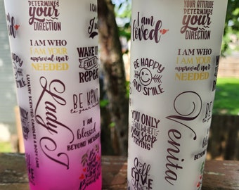 May include: Two white tumblers with motivational quotes and names "Lady T" and "Venita" printed on them. The tumblers have a wooden lid and a pink and white gradient.