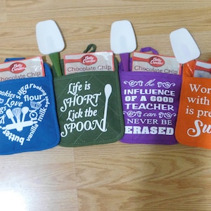 Puede incluir: Cuatro agarraderas de olla coloridas con diferentes refranes. Las agarraderas son verdes, moradas, azules y naranjas. Cada agarradera tiene una espátula blanca unida a ella. Los refranes en las agarraderas son "La vida es corta, lame la cuchara", "La influencia de un buen maestro nunca se puede borrar", "Galletas Azúcar Hornear Amor Harina Huevos Mantequilla Vainilla Leche", y "Trabajar contigo es bastante dulce!"