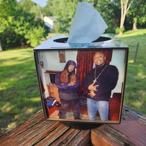 May include: A black and white tissue box with a photo of two people inside. The tissue box is sitting on a wooden surface with a green background of trees and grass.