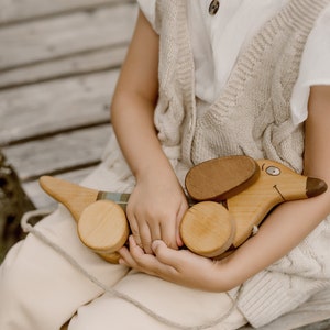 Może przedstawiać: Dziecko trzyma drewnianego psa-zabawkę z brązowymi uszami i namalowaną buzią. Pies ma kółka i jest wykonany z jasnobrązowego drewna. Dziecko ma na sobie białą koszulę i beżową dzianinową kamizelkę.