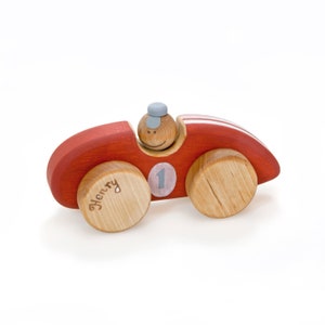 Puede incluir: Un coche de juguete de madera rojo con una figura de madera sonriente en el asiento del conductor. El coche tiene un número "1" blanco en el lateral y el nombre "Henry" está grabado en una de las ruedas.