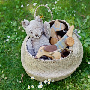Puede incluir: Una cesta de mimbre llena de un oso de peluche gris, un caballo de juguete de madera y un dachshund de juguete de madera. La cesta est&aacute; sentada sobre un lecho de hierba verde con flores blancas.