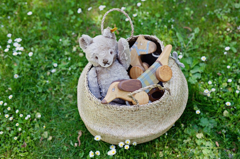 Puede incluir: Una cesta de mimbre llena de un oso de peluche gris, un caballo de juguete de madera y un dachshund de juguete de madera. La cesta est&aacute; sentada sobre un lecho de hierba verde con flores blancas.