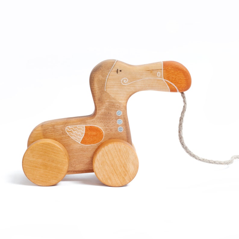 Pu&ograve; includere: Un giocattolo di legno a forma di dodo da tirare, dipinto di marrone, arancione e bianco. Il giocattolo ha due ruote e una corda attaccata al suo becco.