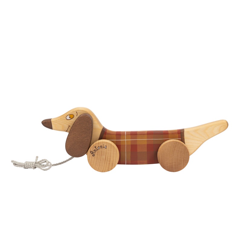 Puede incluir: Un juguete de madera en forma de dachshund con un patr&oacute;n de cuadros rojos y marrones en su cuerpo. El juguete tiene una cuerda para tirar y ruedas, y el nombre "Antonia" est&aacute; escrito en una de las ruedas.