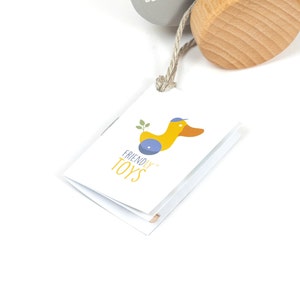 Puede incluir: Una etiqueta blanca con una ilustraci&oacute;n de pato amarillo y el texto "FRIENDLY TOYS" en ella. La etiqueta est&aacute; unida a un juguete de madera.
