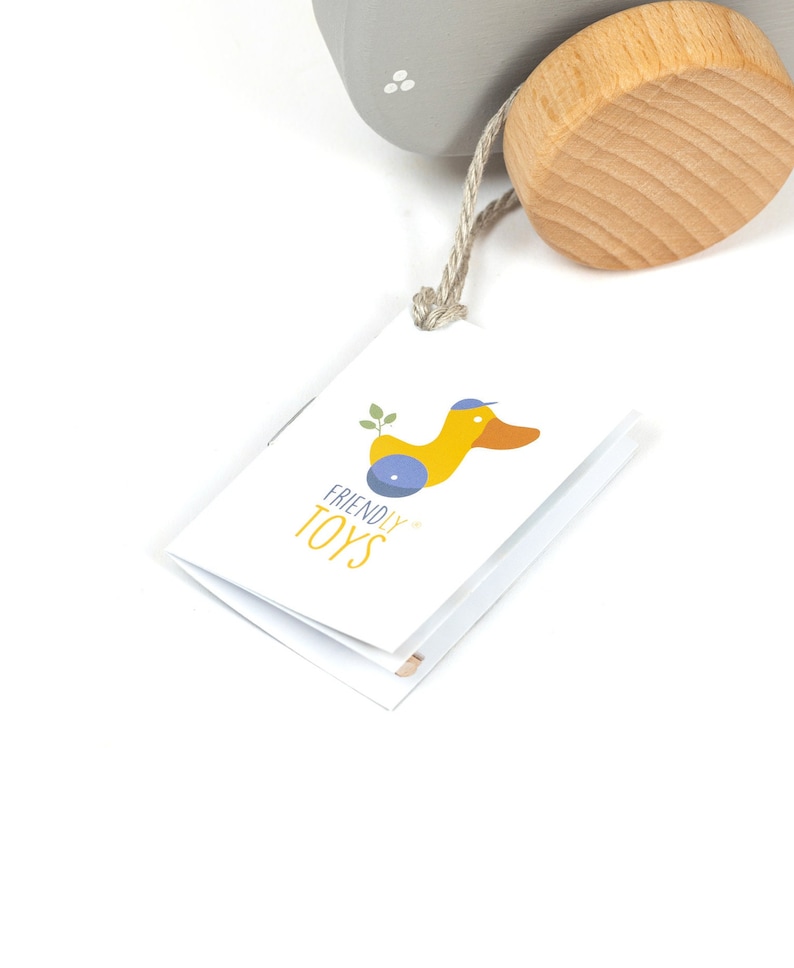 Puede incluir: Una etiqueta blanca con una ilustraci&oacute;n de pato amarillo y el texto "FRIENDLY TOYS" en ella. La etiqueta est&aacute; unida a un juguete de madera.
