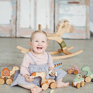 Pu&ograve; includere: Una bambina sorridente gioca con macchinine e animali di legno. I giocattoli sono dipinti in colori pastello e hanno facce di animali. La bambina indossa una maglietta rosa e una salopette blu.