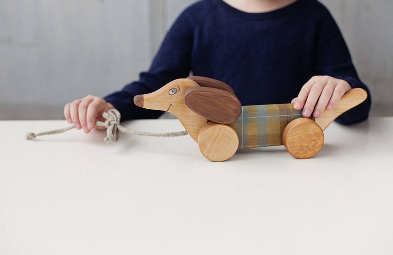 Puede incluir: Un juguete de madera en forma de dachshund con orejas marrones y un cuerpo de tela a cuadros. El juguete tiene ruedas y una cuerda atada.