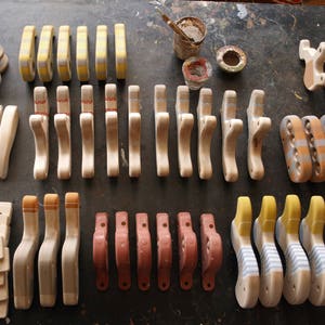 Pu&ograve; includere: Una collezione di ganci in legno con vari disegni dipinti, tra cui strisce, punti e motivi geometrici. I ganci sono disposti in file su una superficie strutturata.