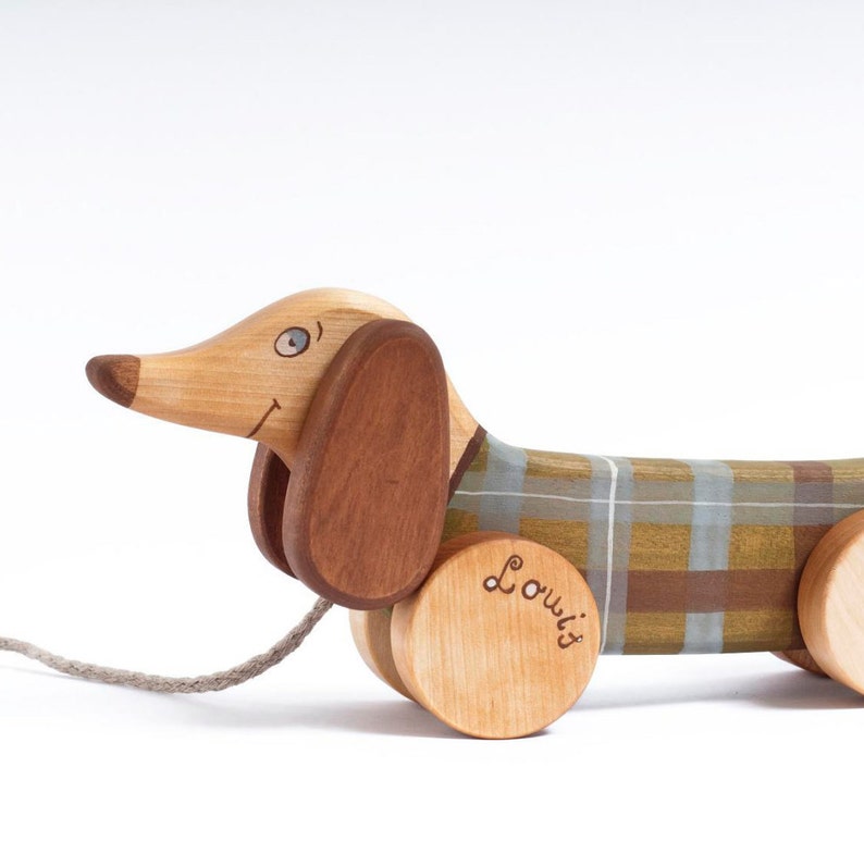 Puede incluir: Un juguete de madera en forma de dachshund con tela de cuadros marr&oacute;n y beige, orejas marrones y un ojo azul. El juguete tiene una cuerda para tirar y ruedas. La palabra "Louwi's" est&aacute; escrita en la rueda.